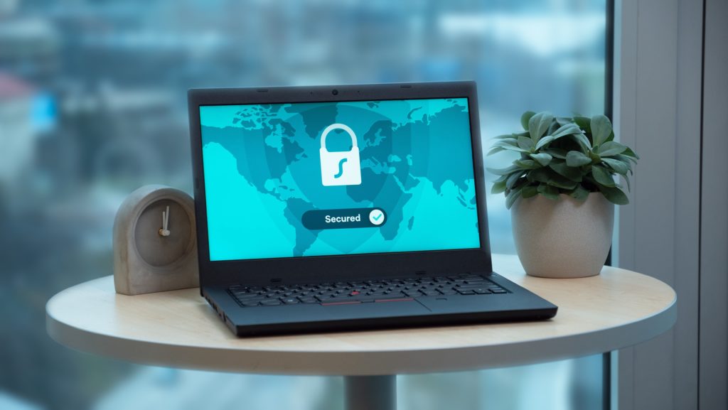 Image of an open laptop on a small round table.  The laptop is displaying a blue world map with a white padlock superimposed over the top and the word secure under it.  To the right of the laptop is a small clock and to the right is a small potted plant.