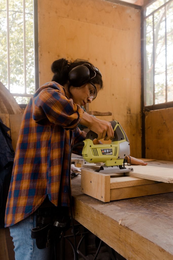 Image of a woman dressed in jeans and an orange and blue flannel shirt using a power tool to assemble two pieces of wood.