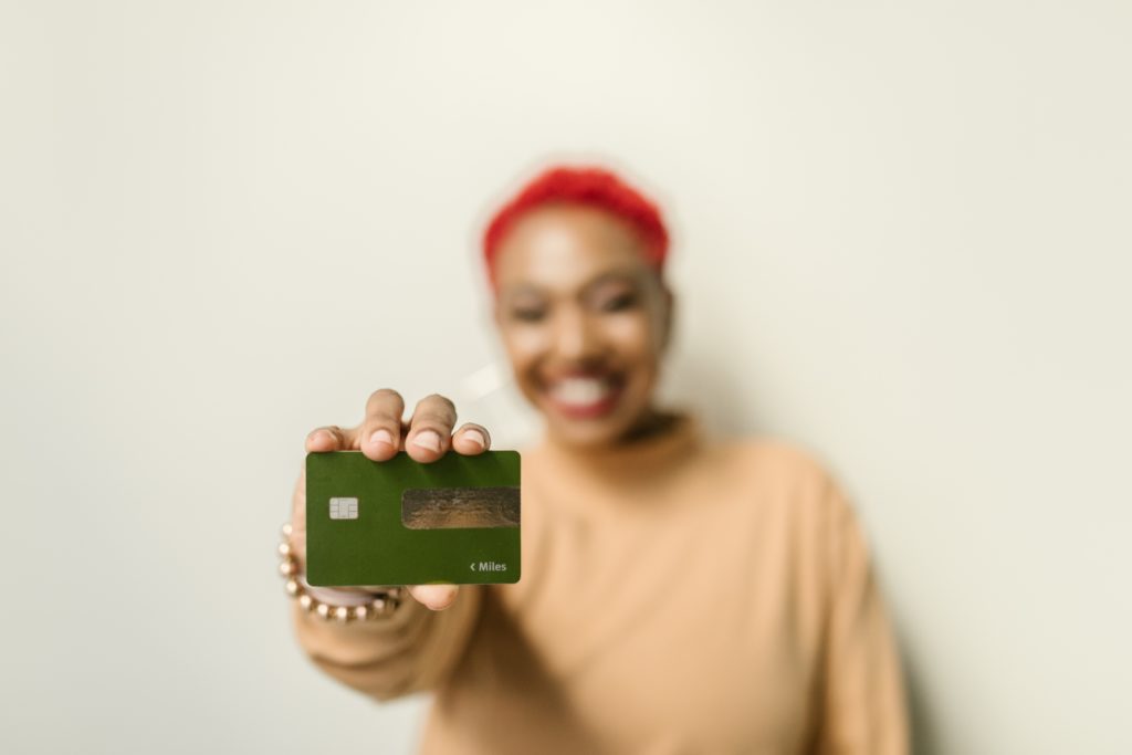African American woman with short red hair dressed in a cream coloured swater holding a green credit card in her outstretched hand