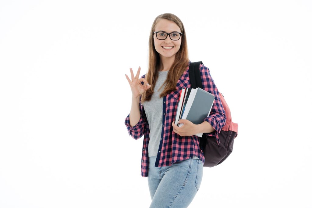 Photo of a young  woman with  long dark blonde hair and black glasses dressed as a typical college student.  She is holding several text books and is wearing a backpack.  She is dressed in blue jeans, grey tshirt and an flannel overshirt.  Her emty hand is sowing the "ok" gesture.