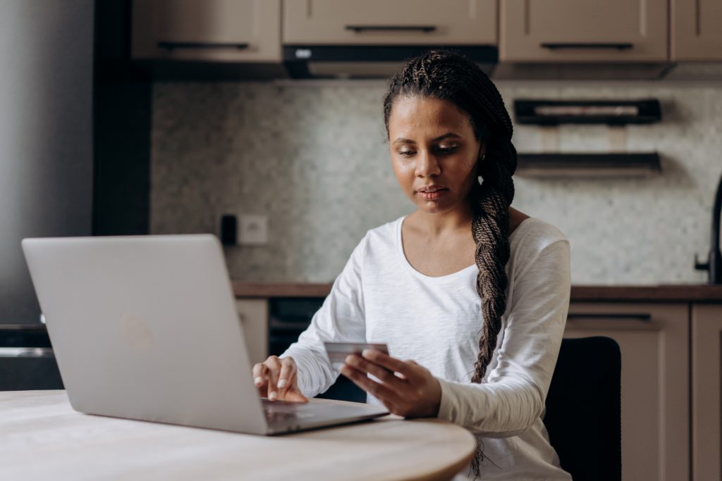 Image of a woman sitting at a kitchen table.  There is an open laptop on the table and she is holding a credit card in her hand.