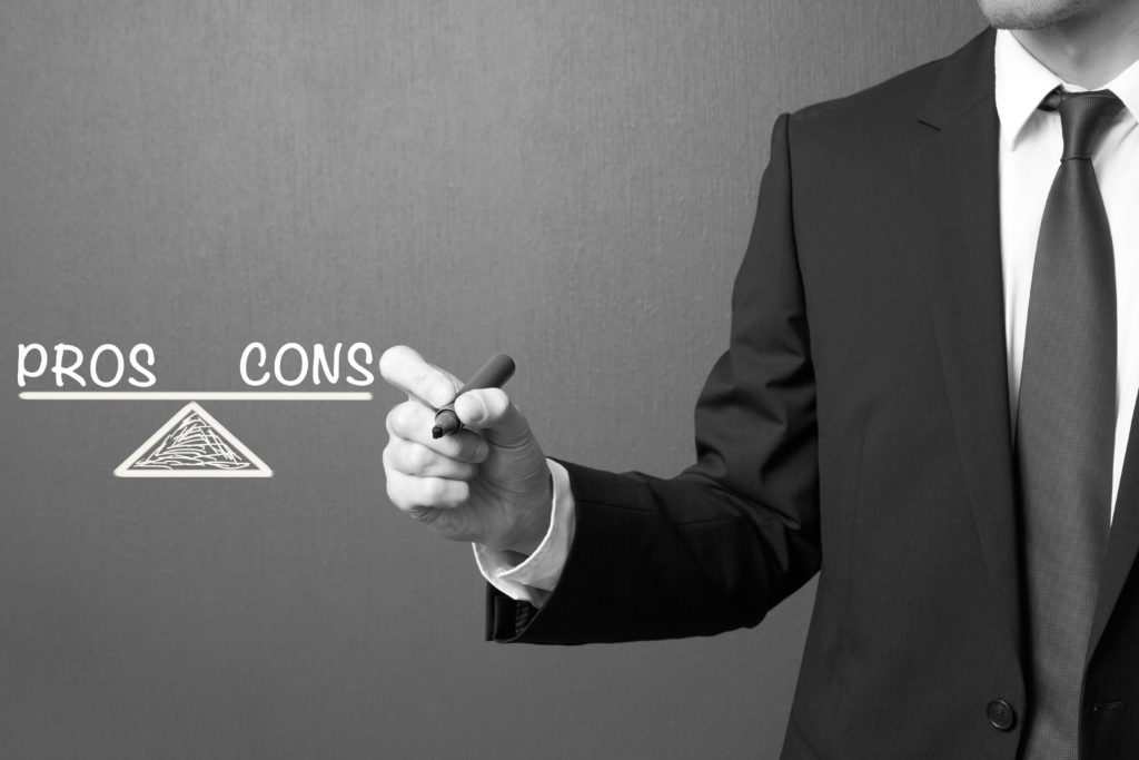 black and white image of a man in a suit pointing to the words pros and cons balanced on a scale