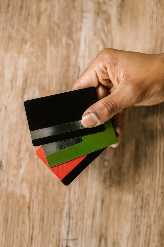 brown skinned hand holding three credit cards in a fan shape,  Top card is black, middle cars is green, and bottom card is red.