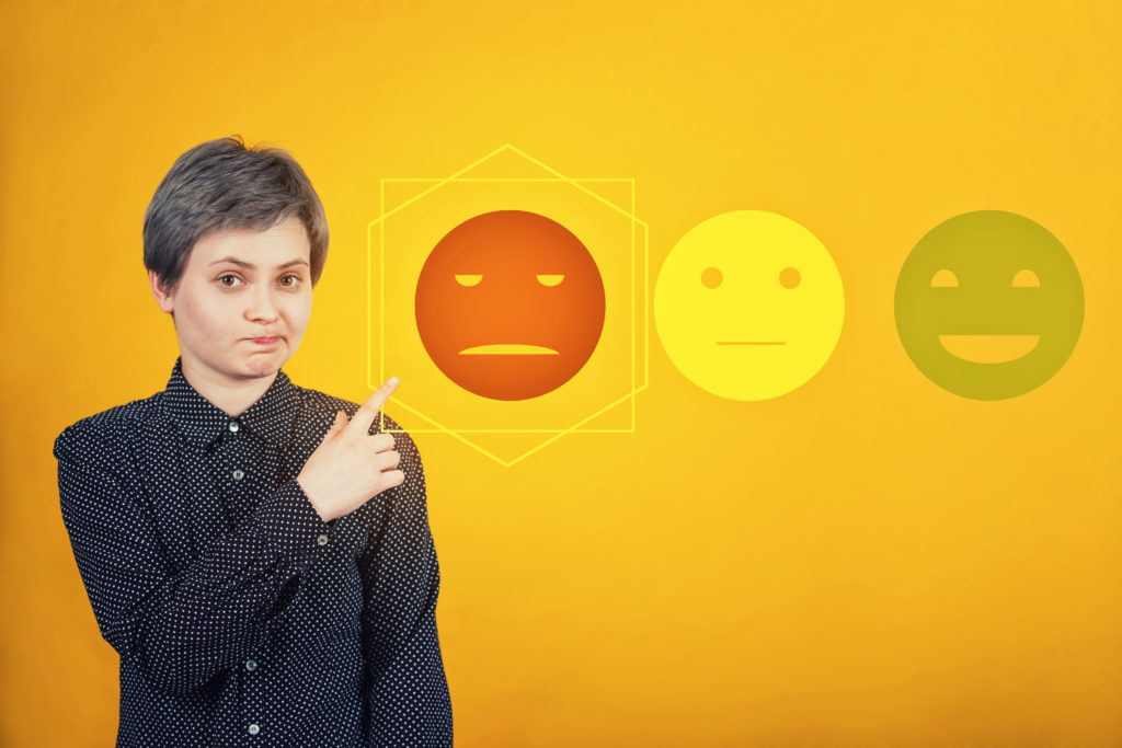woman with a look if disappointment on her face pointing at a drawing of a red sad face next to a yellow neutral face and green happy face.  The woman is wearing a long sleeved black shift and the background is orange