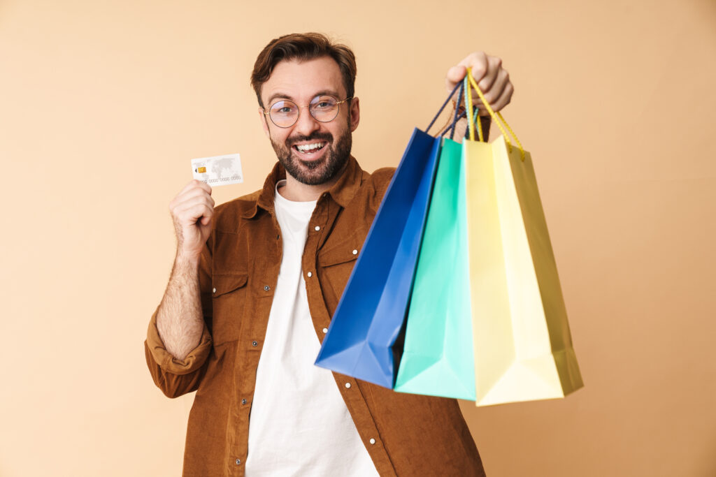 Image of a pleased positive cheery young unshaved man isolated over beige wall background holding credit card and shopping bags.