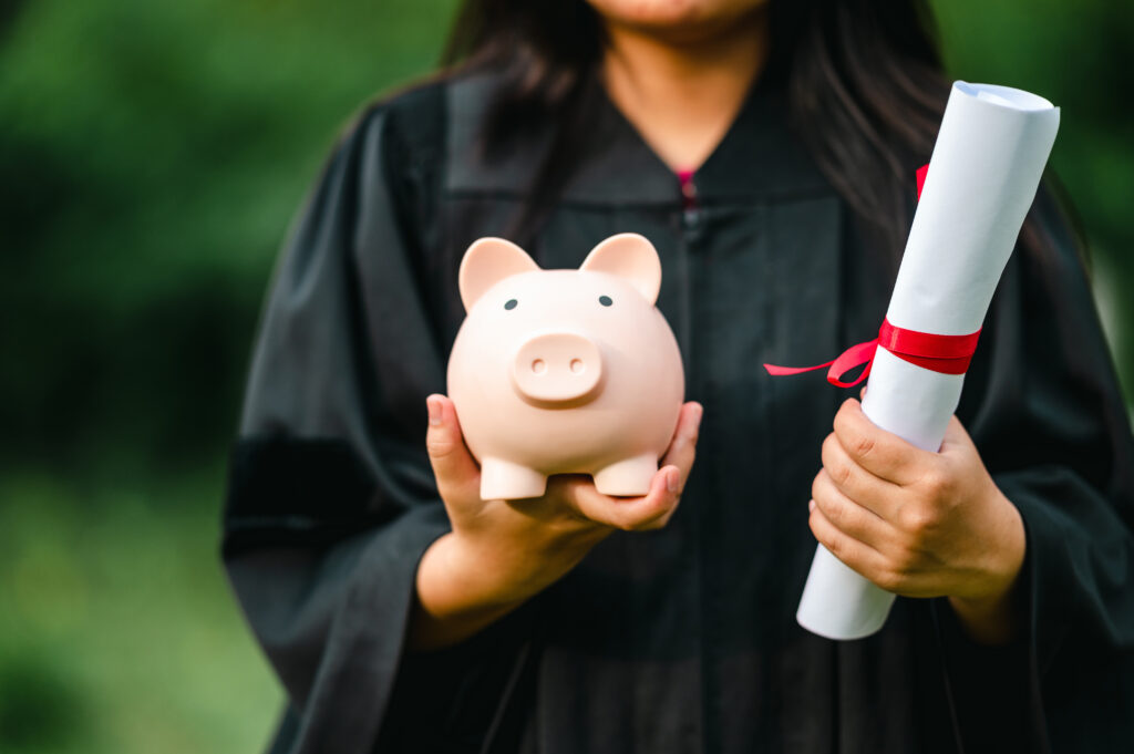 close up of a woman in a black graduation gown holding a diploma scroll in her left hand and a pink piggy bank in her right hand