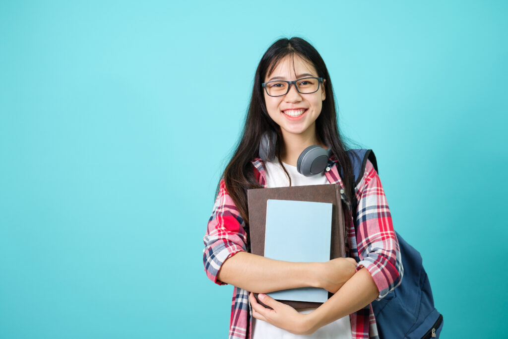 light blue background with smiling young Asian woman wearing a red plaid shirt, headphones around her neck, and a backpack over her right shoulder holding books in her hands.