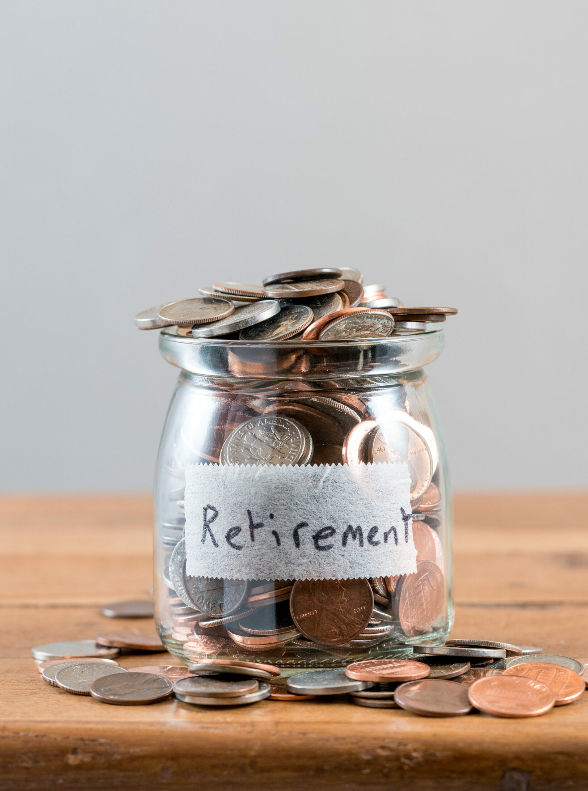 glass jar sitting on a wooden table fill with coins that are spilling over the top.  The jar has a piece of masking tape on it with the word retirement written in black letters