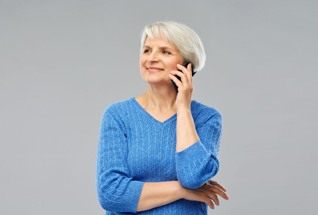 light gray background, older woman with short grey hair wearing a blue shirt holding a black mobile phone to her ear